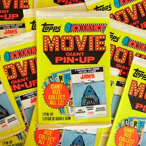 Può includere: Una collezione di carte da collezione Topps Real Movie Giant Pin-Up con il film Lo squalo. Le carte sono gialle, rosse e blu con un'immagine di uno squalo e il testo "Jaws".