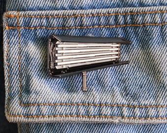 May include: A silver-colored metal pin shaped like a movie studio spotlight with a black base and white light bulbs.