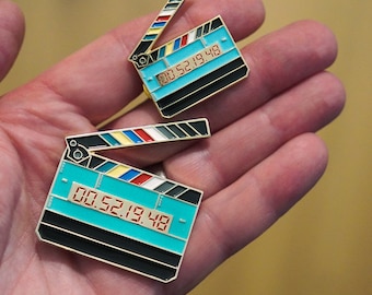 May include: Two gold-colored enamel pins shaped like a clapperboard with a blue background and a red digital display showing "00:52:19:48".