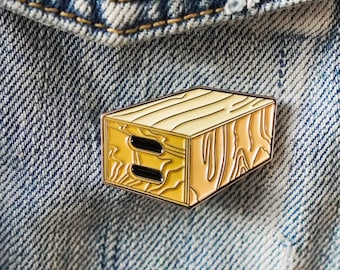 May include: A wooden crate-shaped enamel pin with a light brown wood grain pattern and two black rectangular cutouts. The pin is attached to a denim jacket.