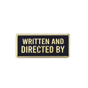 Op de afbeelding: Een rechthoekige pin met de tekst "WRITTEN AND DIRECTED BY" in gouden letters op een zwarte achtergrond. De pin heeft een gouden rand. Deze pin is een knipoog naar filmproductie.