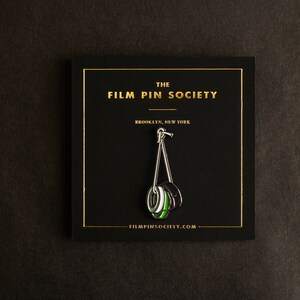 May include: A black and green enamel pin depicting a film reel hanging from a hook. The pin is displayed on a black card with gold lettering that reads "The Film Pin Society, Brooklyn, New York, filmpinsociety.com".