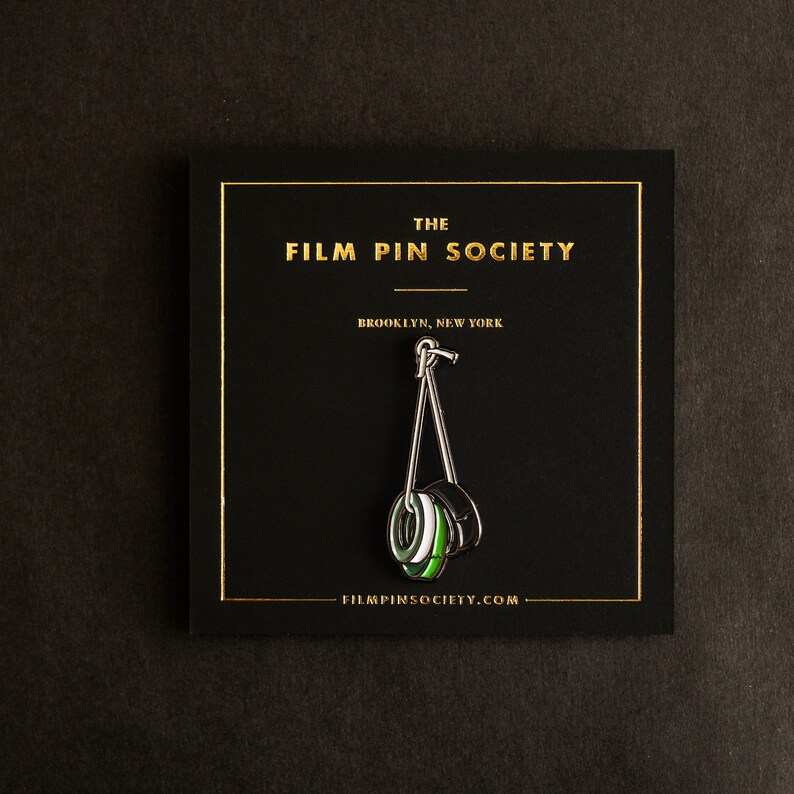 May include: A black and green enamel pin depicting a film reel hanging from a hook. The pin is displayed on a black card with gold lettering that reads "The Film Pin Society, Brooklyn, New York, filmpinsociety.com".