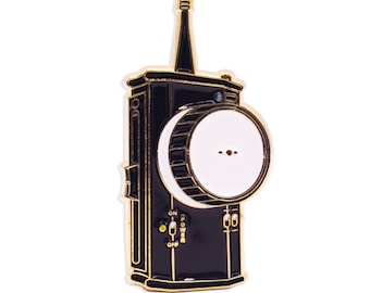 May include: A black and gold enamel pin shaped like a vintage camera with a white lens and a small blue detail. The pin has a gold 'ON' and 'OFF' switch and a gold 'POWER' button.