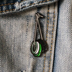 May include: A green, white and black enamel pin of three stacked tyres hanging from a silver chain. The pin is attached to a blue denim jacket.