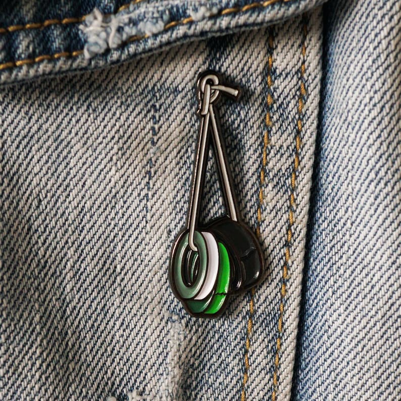 May include: A green, white and black enamel pin of three stacked tyres hanging from a silver chain. The pin is attached to a blue denim jacket.