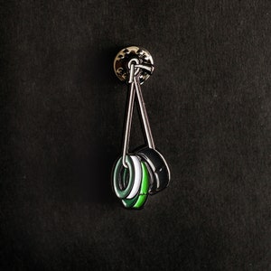 May include: A silver pin with three black and green rings hanging from it. The rings are layered on top of each other and have a white stripe.