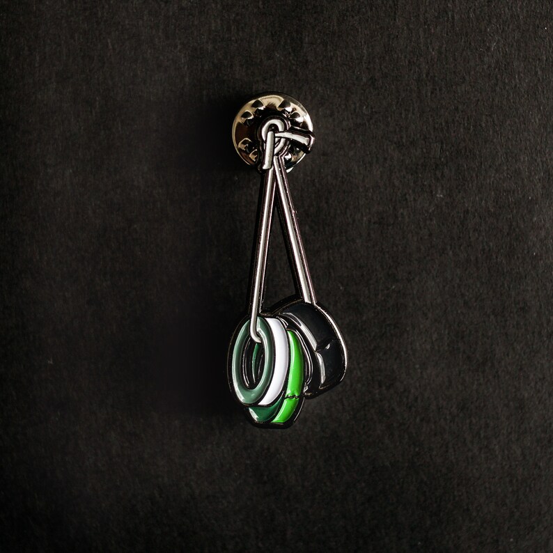May include: A silver pin with three black and green rings hanging from it. The rings are layered on top of each other and have a white stripe.