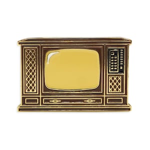Op de afbeelding: Een goudkleurige pin in de vorm van een vintage televisie. De tv heeft een bruine lijst met gouden accenten, een geel scherm en een zwart bedieningspaneel. Het ontwerp bevat luidsprekerachtige details aan de zijkanten, wat een retro-esthetiek oproept.