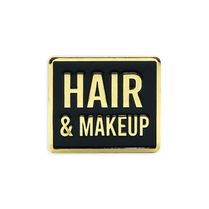 May include: Square pin with a black background and gold trim. The pin features the words "HAIR & MAKEUP" in gold lettering. The pin is a beauty-themed accessory.
