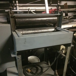May include: A vintage industrial machine with two large, cylindrical rollers. The machine is painted a light gray and sits on a metal frame with a lower shelf. A black power cord with a yellow plug is coiled on the shelf.