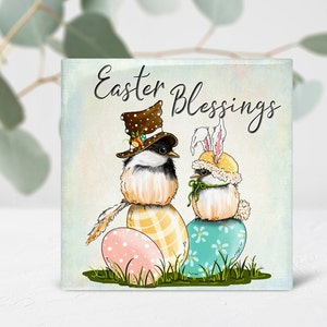 May include: Square Easter-themed artwork featuring two birds perched on decorated Easter eggs. One bird wears a top hat, the other a bunny ear bonnet. The artwork includes the text "Easter Blessings" in script lettering. The background is a soft, pastel watercolor.