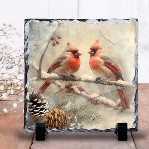 May include: A square slate tile featuring a winter scene with two red cardinals perched on a snow-covered branch. Pine cones and berries are also visible. The tile is supported by a black stand.