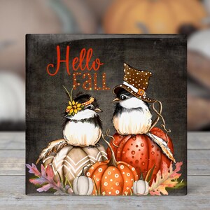 May include: A square decorative sign with a dark gray background featuring the text Hello Fall in orange. Two birds sit on pumpkins; one wears a sunflower headband, the other a brown polka-dot hat. The scene includes orange and white pumpkins and autumn leaves.