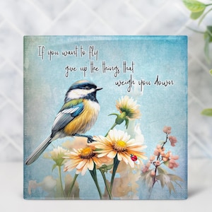 May include: Square wall art featuring a colorful bird perched on yellow and white flowers. The artwork has a light blue background with the text "If you want to fly give up the things that weigh you down."