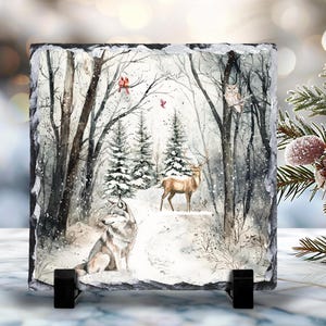 May include: A square decorative slate tile featuring a winter forest scene. The artwork depicts a wolf, deer, and owl in a snowy landscape with trees and falling snow. The tile is supported by a black stand.