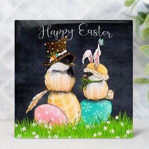 May include: Square Easter-themed artwork featuring two whimsical birds. One bird wears a polka-dot top hat, the other a bunny ear hat. They sit atop decorated eggs, with the words "Happy Easter" in script above.