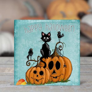 May include: A teal blue card with the text "Happy Halloween" and a black cat sitting on top of two pumpkins. The pumpkins are orange and have carved faces. There are also two small black birds on the card.