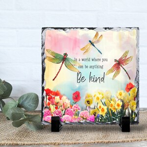 May include: A square slate plaque with a watercolor background of pink, blue, and yellow. Three dragonflies are flying over a field of yellow daffodils, pink and red tulips, and a single red poppy. The text "In a world where you can be anything Be kind" is printed on the plaque.
