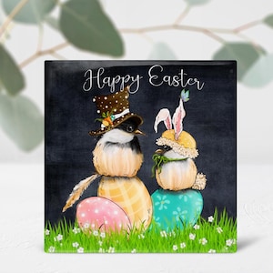 May include: Happy Easter greeting card with a black background. Two birds wearing hats and holding Easter eggs are depicted. The bird on the left is wearing a black and white top hat with a green feather. The bird on the right is wearing a yellow bunny hat with pink ears. Both birds are holding colorful Easter eggs.