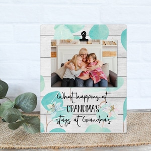 May include: A white wood picture frame with a light blue watercolor floral design. The frame has a clip at the top and a photo of a grandmother and two grandchildren. The text on the frame reads "What happens at Grandma's stays at Grandma's".