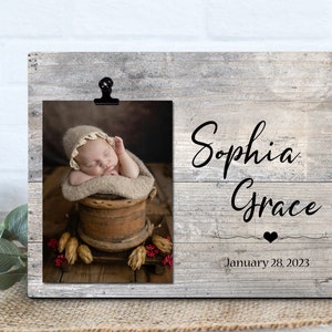 May include: A rustic wooden photo frame with a clip to hold a picture. The frame has a gray woodgrain finish and the text "Sophia Grace" and "January 28, 2023" is printed on it. The photo shows a baby sleeping in a wooden bucket.