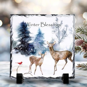May include: A slate coaster with a winter scene featuring two deer in a snowy forest. The text "Winter Blessings" is printed on the coaster.