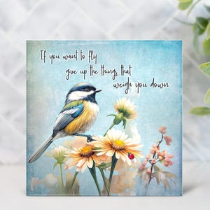 May include: Square canvas art featuring a colorful bird perched on yellow and white flowers. The artwork has a light blue background with the quote, "If you want to fly give up the things that weigh you down."