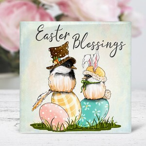 May include: Easter Blessings card featuring two cartoon birds wearing hats. One bird is wearing a brown top hat with a carrot and the other is wearing a yellow bunny hat with bunny ears. Both birds are sitting on colorful Easter eggs.