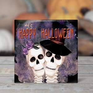 May include: A square, purple and black Halloween-themed coaster with a watercolor design of two skeletons wearing hats and flowers. The text "Happy Halloween" is printed in orange on the coaster.