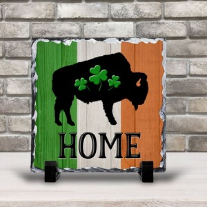 May include: A square slate sign featuring a black bison silhouette with three green shamrocks, set against a backdrop of the Irish flag. The word "HOME" is displayed in bold, black letters. The sign is supported by a black stand.