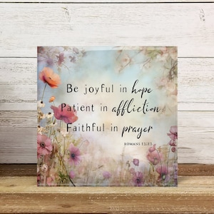 May include: Square decorative art piece with floral and botanical design. The artwork features the text "Be joyful in hope, Patient in affliction, Faithful in prayer" from Romans 12:12. The background has a soft, watercolor-like effect with pink and orange flowers.