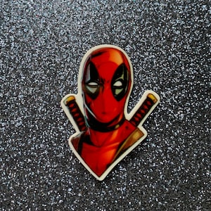 May include: A red and black Deadpool character sticker with a white background. The sticker features Deadpool's head and shoulders with his signature swords.