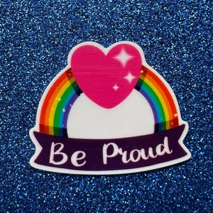 May include: A rainbow with a pink heart on top and a white banner with the text "Be Proud" in purple lettering.