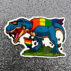 May include: A blue dinosaur wearing sunglasses and a rainbow cape holds a rainbow flag. The dinosaur is standing on a green patch of grass with leaves scattered around it.