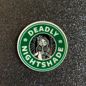 Puede incluir: Una pegatina redonda verde y blanca con el texto "Deadly Nightshade" en un c&iacute;rculo alrededor de una imagen de dibujos animados de una mujer con el pelo largo y negro y un vestido blanco.