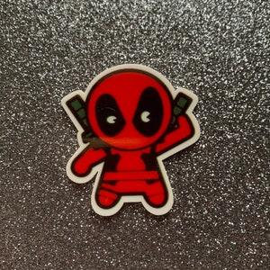 May include: A red and black cartoon character sticker of Deadpool with a white background. The character is in a fighting pose with his arms raised.