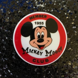 Peut inclure: Un badge rond du Mickey Mouse Club avec une bordure rouge et blanche. Il présente le visage de Mickey Mouse et les mots "MEMBER 1955" et "Mickey Mouse Club". Le fond est une surface sombre et scintillante.