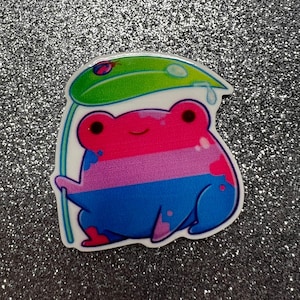 May include: A cartoon frog with a pink, blue, and white striped body is holding a green leaf with a raindrop. The frog is smiling and has a happy expression.