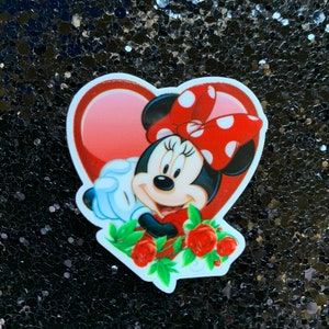 Peut inclure: Un autocollant en forme de cœur rouge avec une image de dessin animé de Minnie Mouse portant un nœud rouge à pois blancs. Elle tient un bouquet de roses rouges avec des feuilles vertes.