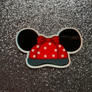Peut inclure: Un chapeau rouge à pois blancs avec des oreilles noires et un nœud rouge. Le chapeau est sur un fond de paillettes argentées.