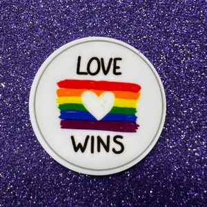 May include: White round sticker with a rainbow flag and a white heart in the center. The text "LOVE WINS" is printed in black below the flag.