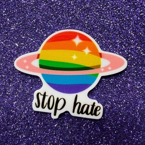 May include: A rainbow-colored planet with a pink ring around it. The planet has white stars on it. The text "stop hate" is written below the planet.
