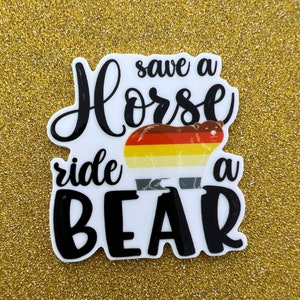 May include: A white sticker with black text that reads "save a horse ride a bear". The sticker features a cartoon bear with a rainbow coloured stripe across its body.