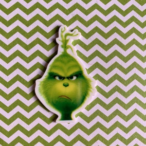 May include: A green Grinch character with a grumpy expression, wearing a red Santa hat, against a white background.