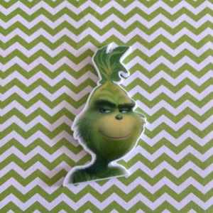 May include: A green Grinch character from the film "The Grinch" with a mischievous expression. The character is depicted in a cartoon style with a large head and a wide smile.