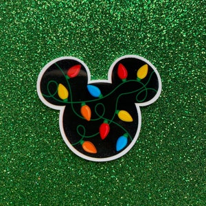 May include: Black and white sticker of a Mickey Mouse head silhouette with colorful Christmas lights wrapped around it. The background is green glitter.