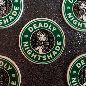 Puede incluir: Cuatro pegatinas redondas verdes y blancas con el texto "Deadly Nightshade" y una imagen de dibujos animados de una ni&ntilde;a con cabello negro y un vestido blanco. Las pegatinas tienen un fondo verde con un dise&ntilde;o de telara&ntilde;a negra y un gr&aacute;fico de calabaza.