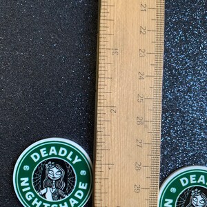 Puede incluir: Dos pegatinas redondas verdes y blancas con una ilustraci&oacute;n en blanco y negro de un personaje con un dise&ntilde;o de telara&ntilde;a y el texto "DEADLY NIGHTSHADE".