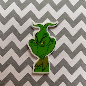 May include: Green Grinch character with a mischievous expression, wearing a green fur collar. The character is depicted in a cartoon style with a pointed hat and large eyes.